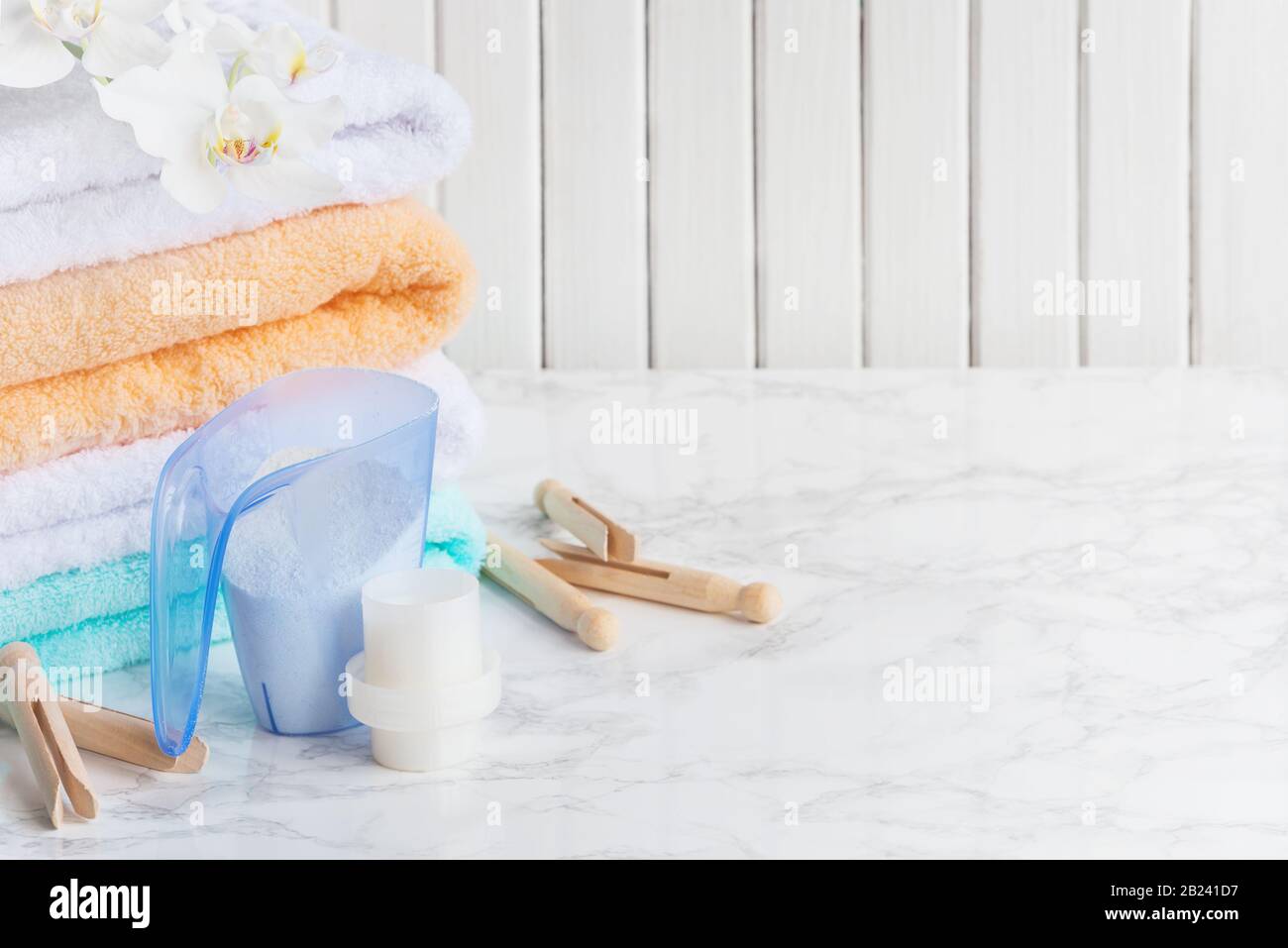 Blaues Kunststoff-Messgefäß mit Waschpulver, Behälter mit Waschmittel, mehrfarbige Frottiertücher, Holzkleiderinnen und weiße Orchidee Stockfoto