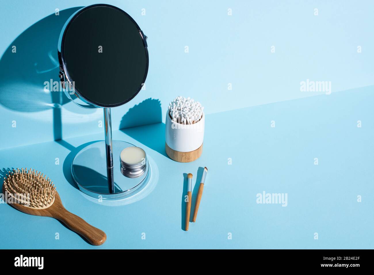 Spiegel mit Wachsbecher neben Haarbürste, Kosmetikbürsten, Zahnbürstenhalter mit Ohrstöpseln auf blauem Grund, Entsorgungskonzept Null Stockfoto