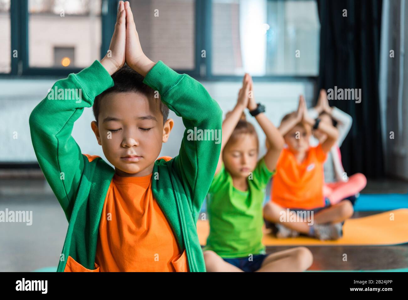 Selektiver Fokus multikultureller Kinder, die mit geklonten Händen über den Köpfen in der Sporthalle meditieren Stockfoto