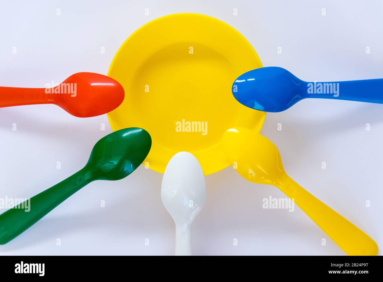 Mehrfarbige Kunststoff-Löffel befinden sich um die Platte auf dem weißen Hintergrund. Flaches Lay. Draufsicht. Stockfoto