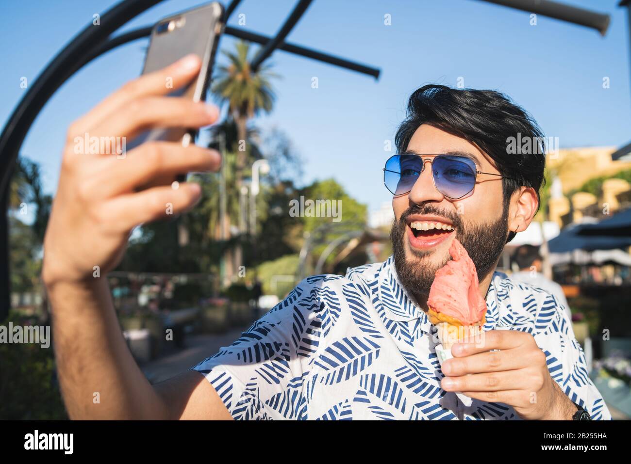 Porträt des jungen Mannes, der sonniges Wetter genießt, mit dem Telefon einen selfie nimmt und draußen eine Eiscreme isst. Stockfoto