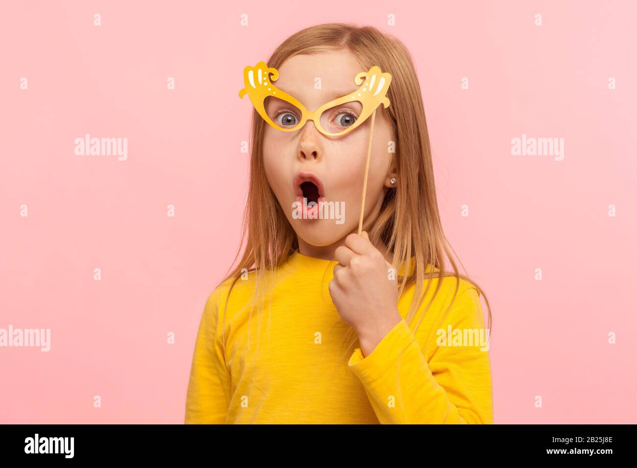 Überraschtes kleines Mädchen, das die Augen mit einer Papierbrille bedeckt und die Zunge herausragt, frech ein Kind, das Spaß hat, eine Maskenmaske trägt und schockiert aussieht. Stockfoto