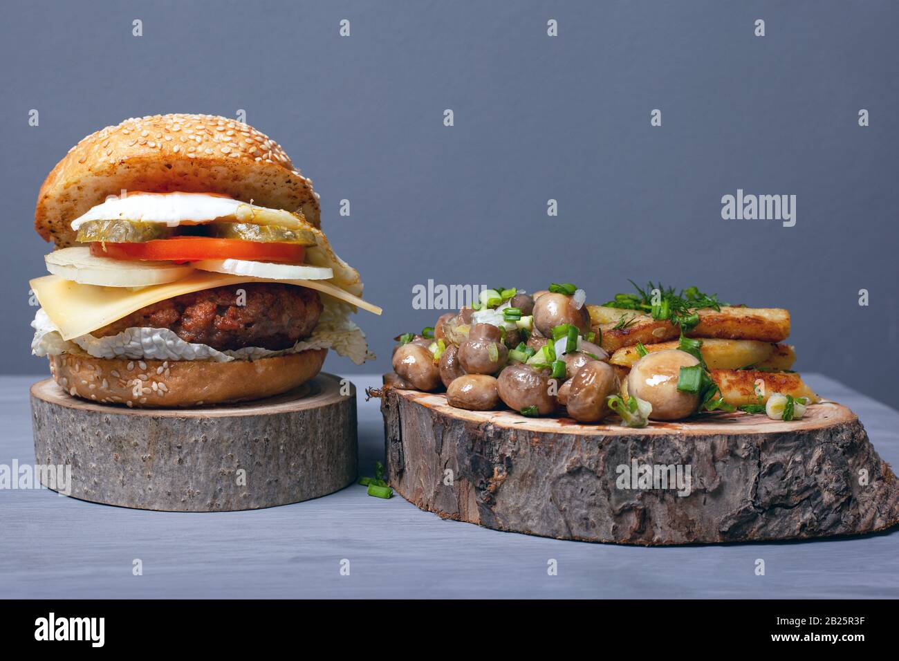 Saftiger Fettburger, gebratene Kartoffeln und Champignons mit Kräutern auf Holztabletts auf grauem Grund, interessante und ungewöhnliche Gerichte für fast Food. Stockfoto