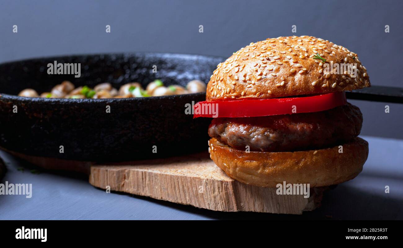 Burger mit einem saftigen Zartbun und Champignonpilzen in einer Kastironpfanne auf einer rustikalen Holzplatte auf grauem Grund. Herzhaftes Mittagessen im Nahbereich. Stockfoto