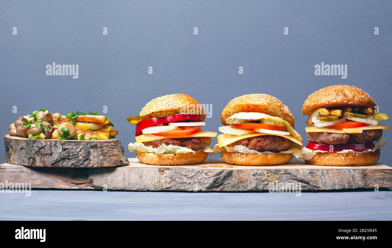 Drei leckere Hamburger mit Pommes und Champignons, die mit grünen Zwiebeln auf grauen Holzküsten bestreut werden. Herzhaftes Mittagessen. Stockfoto