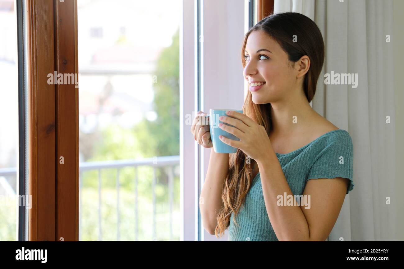 Portrait einer lächelnden Frau, die zu Hause durch das Fenster trinkt und dabei Tee trinkt, Schwerpunkt auf Modellaugen, Innenfoto Stockfoto
