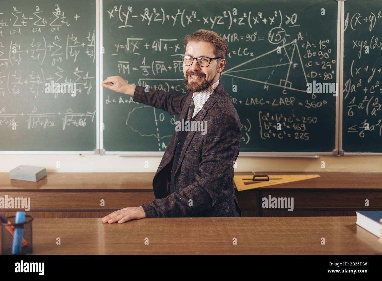 Fröhlicher bärtiger Professor in Brille, der die Formeln erklärt, die er auf einem Schwarzen Brett geschrieben hat Stockfoto
