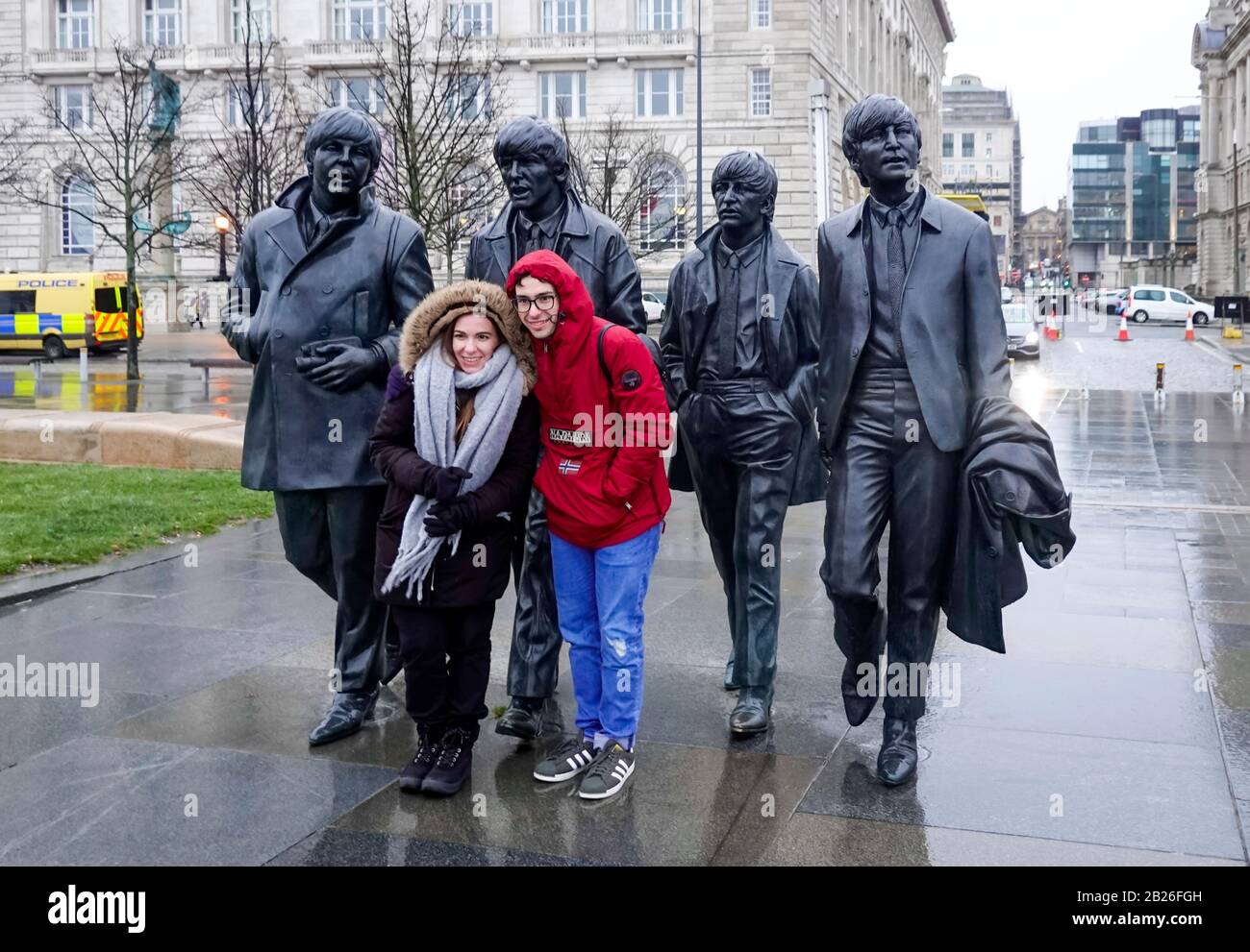 Zwei junge Touristen haben sich gegen den kalten Regen gewickelt und einen Schnappschuss mit der Beatles-Statue in Pierhead in Liverpool gemacht Stockfoto