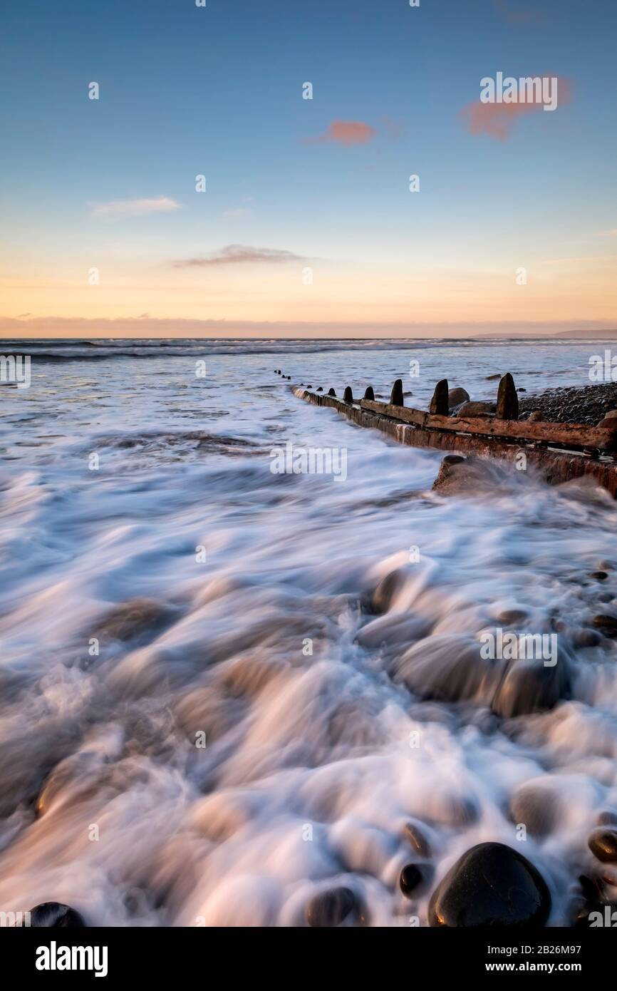 Die Flut, die auf Westward Ho um die Kieselsteine und die Holzmeerschutz-Gabionen spritzt! Strand mit Sonnenuntergang am Horizont, Nord-Devon, Süd-West, Stockfoto