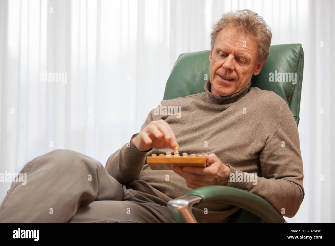 Reifer Mann, der sich in einem Stuhl entspannt, während er in einem hellen Wohnzimmer Solitär spielt Stockfoto
