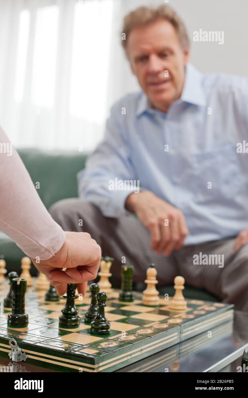 Reifer Mann, der in einem hellen Wohnzimmer Schach spielt - im Vordergrund die Hand der Frau Stockfoto