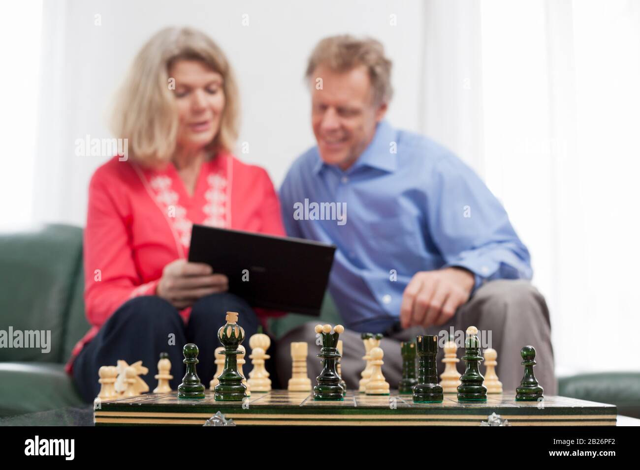Reifes lächelndes Paar mit Blick auf ein Tablet in einem hellen Wohnzimmer - im Vordergrund stehen die Schachleute Stockfoto