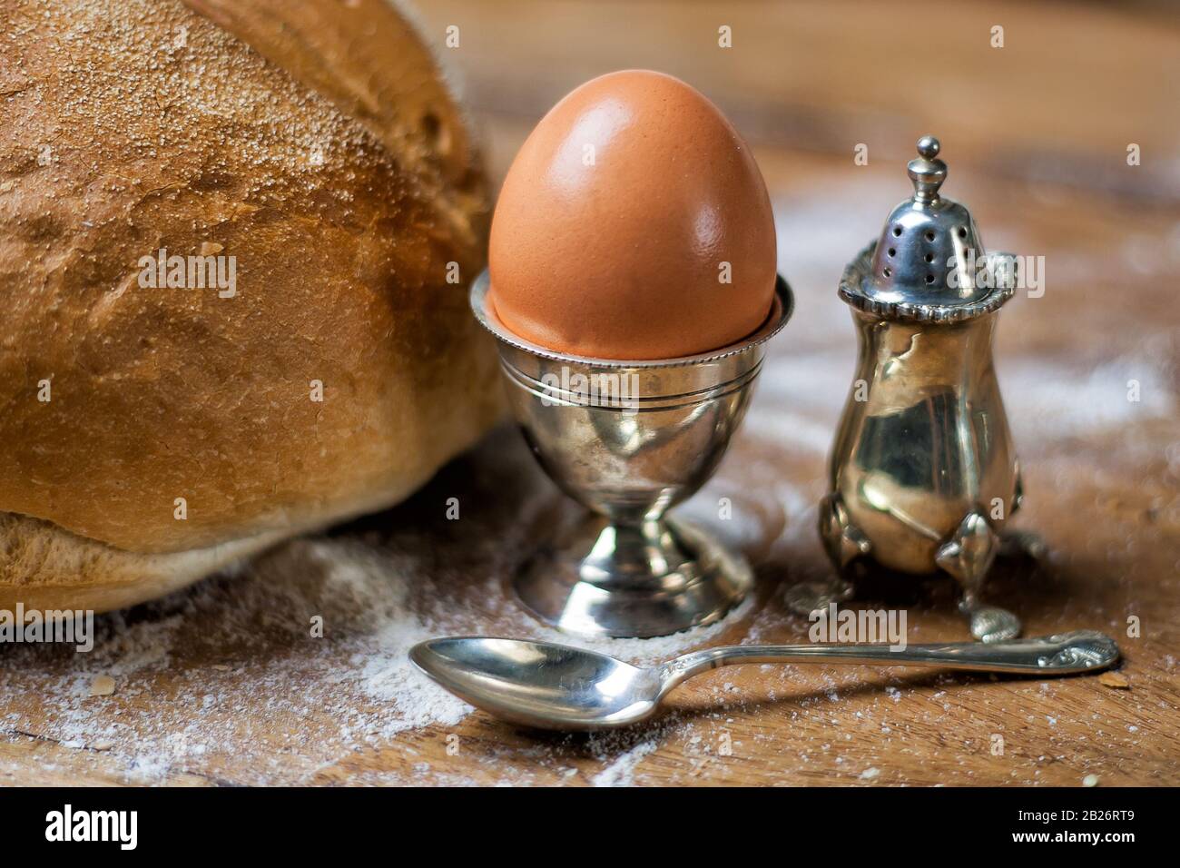 Brot auf dem Holztisch mit Mehl, Ei in silberfarbenem Vintage-Utensil, Salzschüttler und Löffel. Stockfoto