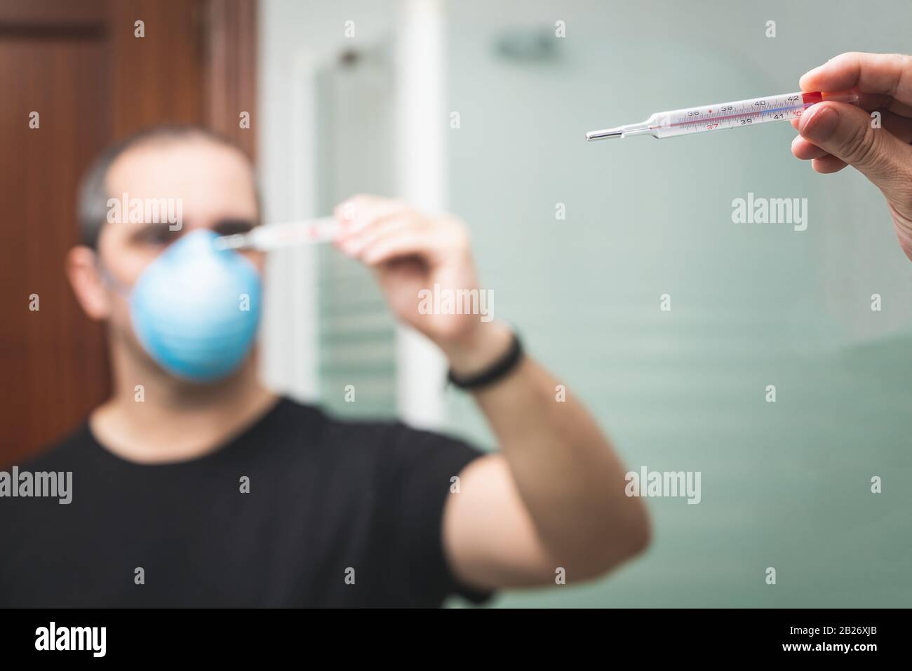 Verschwommener Mann mit medizinischer Maske, das Thermometer sieht Stockfoto
