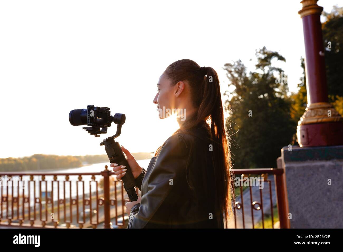 Attraktive junge Brunette mit Kamera-Stabilisator und einem Film im Freien am Morgen. Die Sonne steigt über den Fluss, das sommerliche warme Wetter und Stockfoto