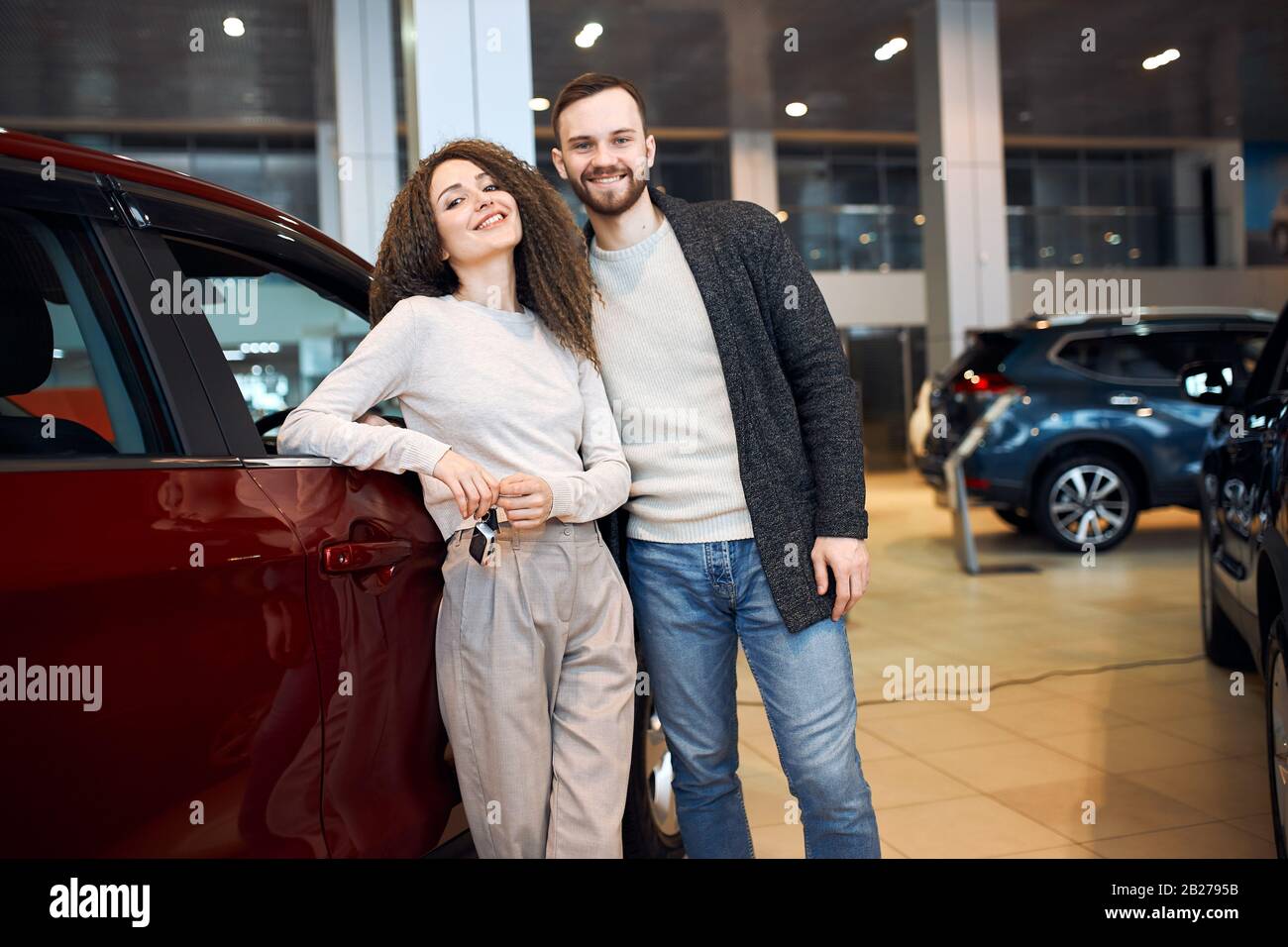 Ehrfürchtiges junges Paar kehrt zurück, um einen Hübschen jungen bärtigen lächelnden Mann zu reisen, der seine schöne Freundin im Autohaus umarmt, die glückliche Frau lächelnd Stockfoto