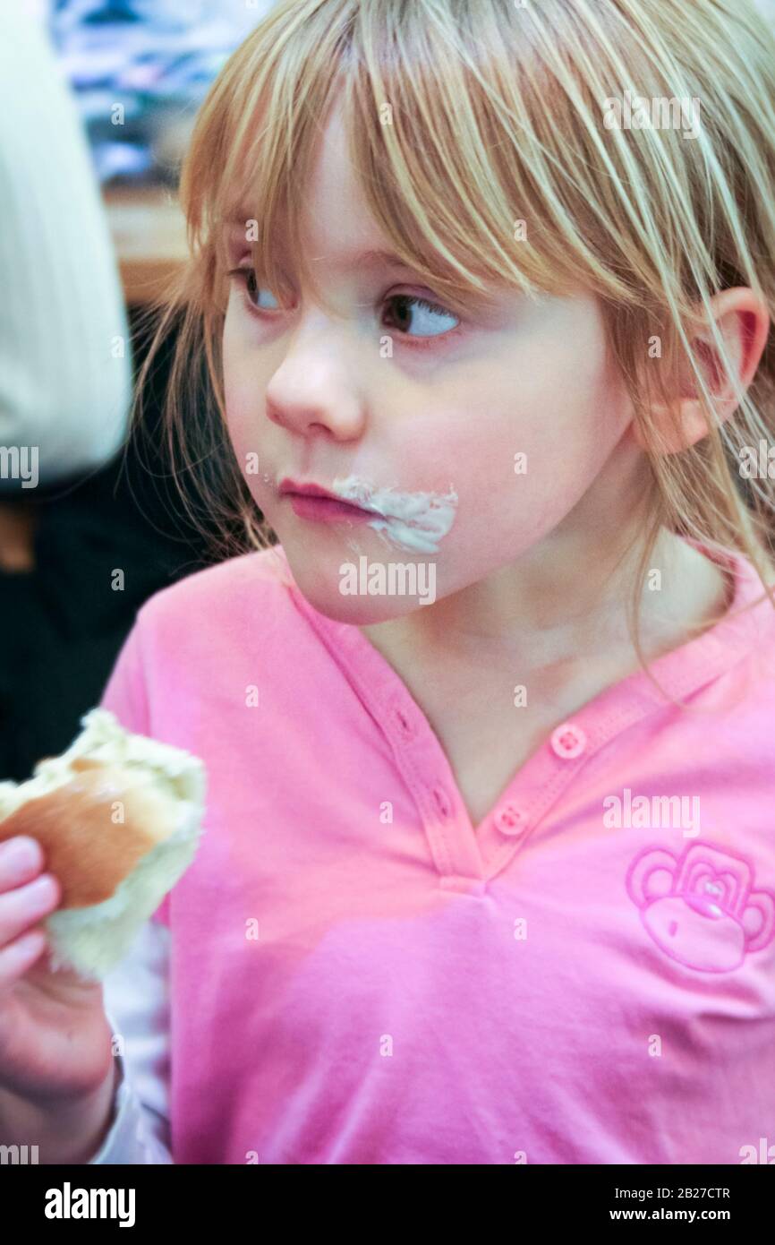 Süßes fünf- oder sechsjähriges Mädchen in einem pinkfarbenen Oberteil, das einen Kuchen mit Sahne um den Mund isst Stockfoto
