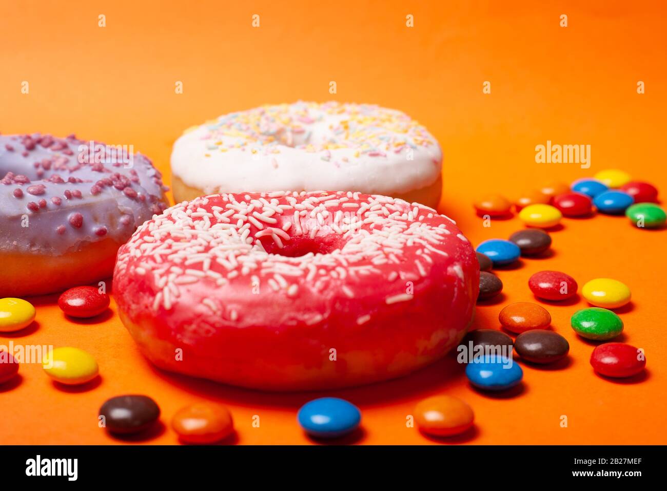 Drei Donuts mit bunter, rot-weißer und violetter Farbe mit eingestreuten Pralinen auf orangefarbenem Hintergrund, Weihnachtsessen Süßigkeiten aus nächster Nähe. Stockfoto