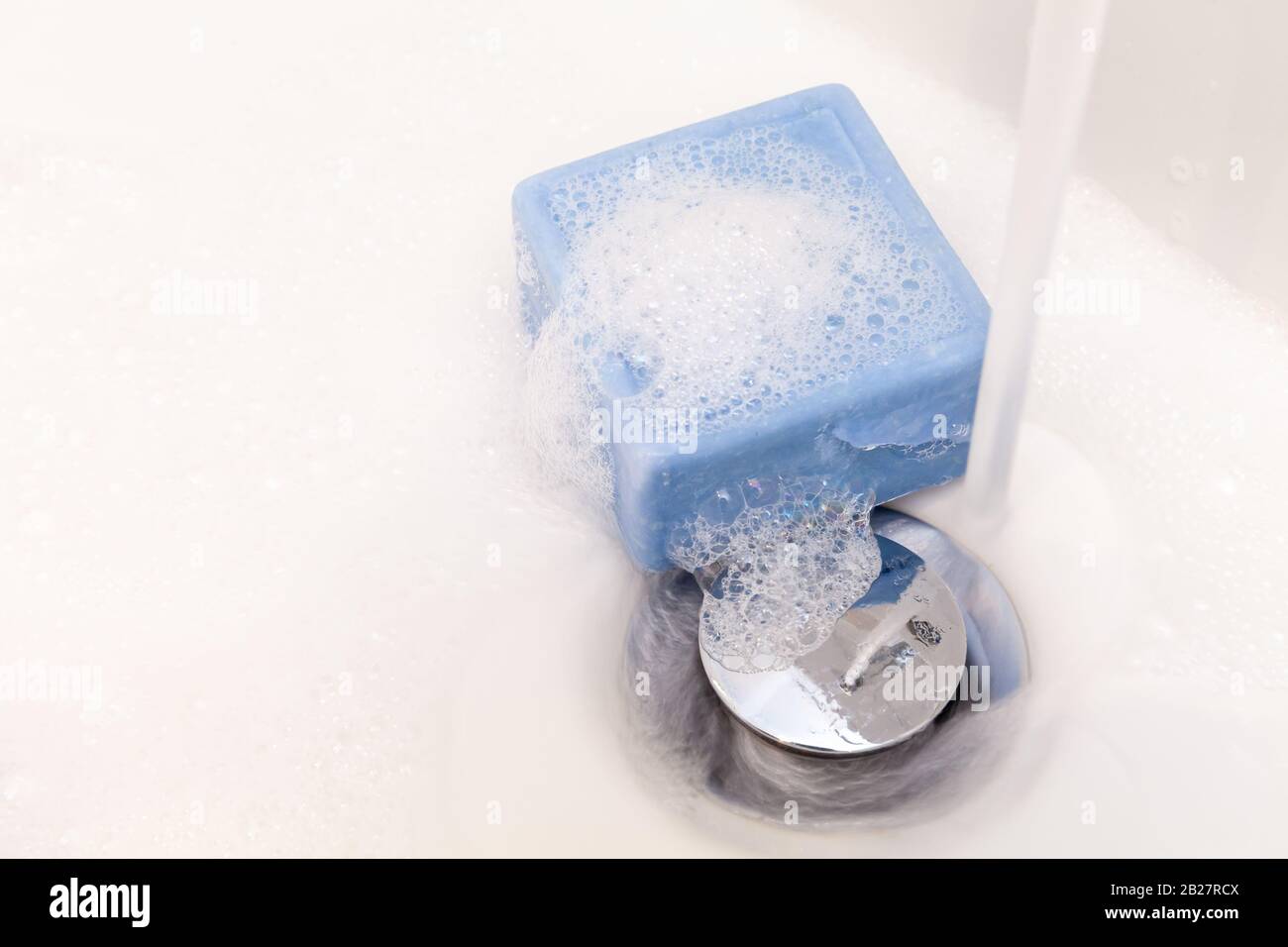 Balken aus quadratischer blauer Seife und saugt in einem weißen Waschbecken mit Chromstecker und fließendem Wasserhahn. Kopierbereich Stockfoto
