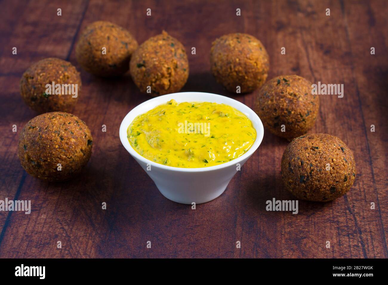 Tahinisauce aus Kurkumulver, Knoblauch und gehackten Petersilienblättern in einer weißen Schüssel. Veganer Kichererbsenfalafel um die Schüssel gelegt. Stockfoto