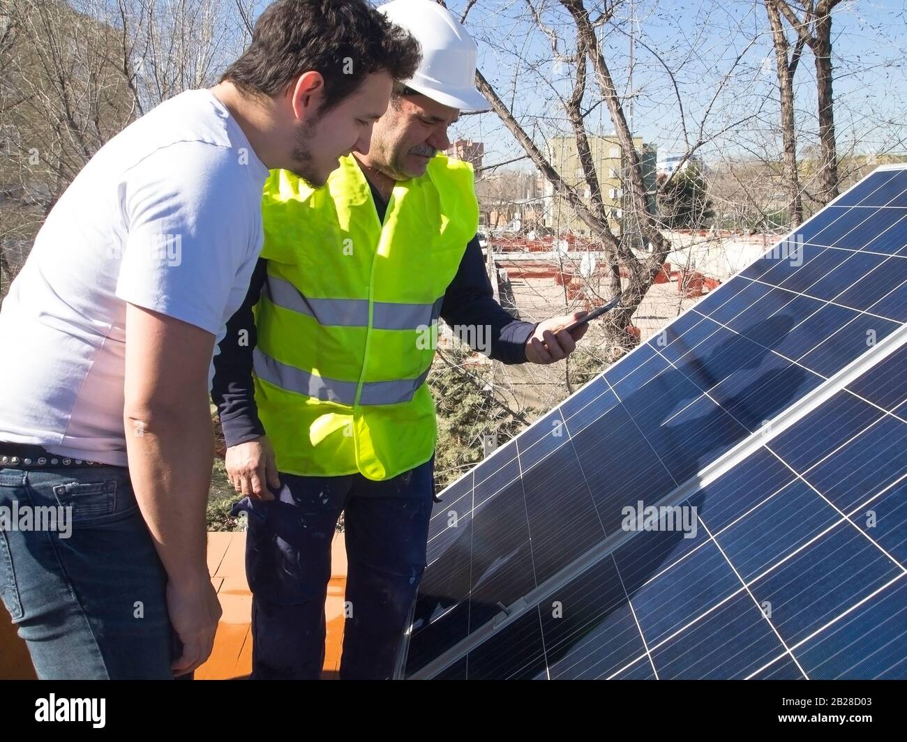 Kaukasischer erfahrener Techniker spricht die App mit seinem jungen Kunden neben den Solarmodulen. Alternative Stromquelle, nachhaltige Ressource Stockfoto