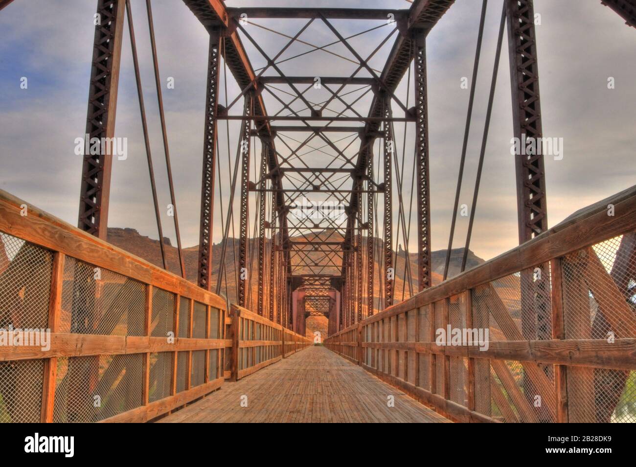 Perspektive Blick auf eine Brücke und ihren Holzdecksteg mit eingezäunten Handläufen aus Holz unter den metallenen Tragwerken in warmer, rostfarbiger Farbe Stockfoto
