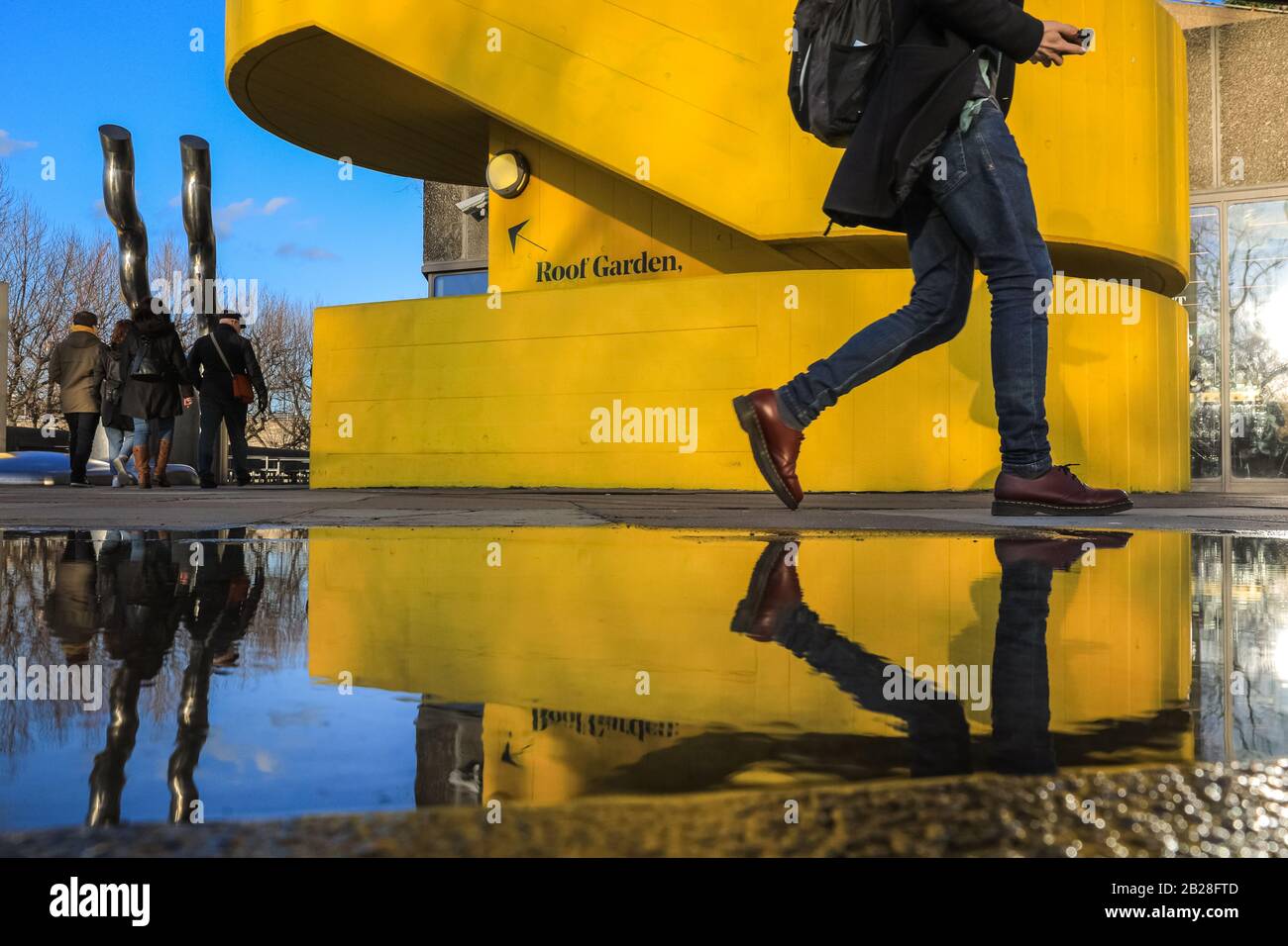 London, Großbritannien. März 2020. Besucher des Londoner Southbank Center spiegeln sich in Regenpfützen wider, wenn sie an einem der berühmten, leuchtend gelben, brutalistischen Architekturtreppen des Zentrums vorbeigehen. Der Tag in London erlebte einen schönen Sonnenschein mit blauem Himmel, in krassem Gegensatz zu den Regenfällen und dem Sturm am Samstag. Kredit: Imageplotter/Alamy Live News Stockfoto