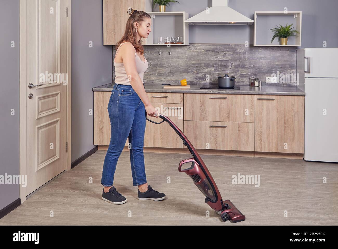 Eine Putzfrau saugt den Küchenboden mit einem elektrischen Besen. Kaukasische Frau, die mit einem schnurlosen, aufrechten Vakuum im Kochraum auftidst Stockfoto