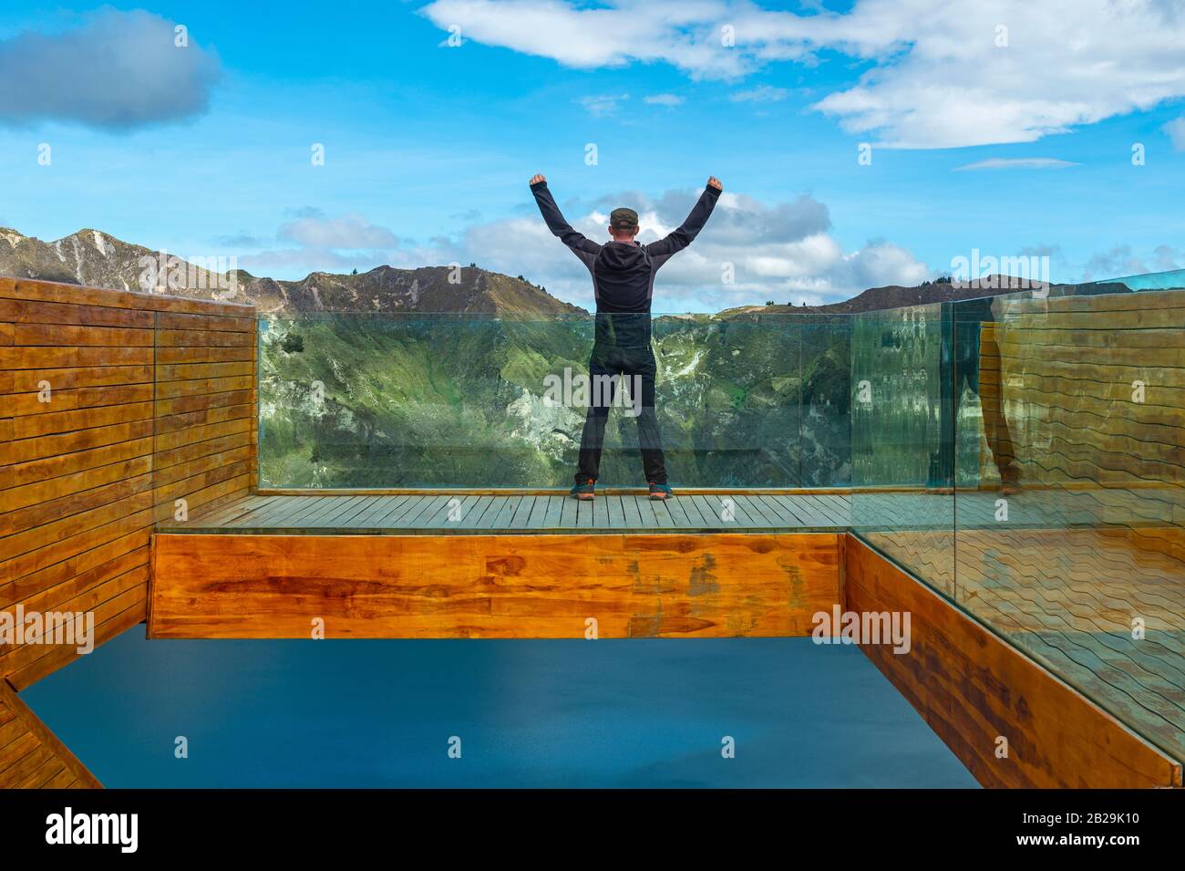 Junger männlicher Rucksacktourist mit ausgestreckten Armen am Himmel auf dem Aussichtspunkt am türkisfarbenen Quilotoa-Kratersee, Quilotoa-Schleifenwanderung, in der Nähe von Quito, Ecuador. Stockfoto