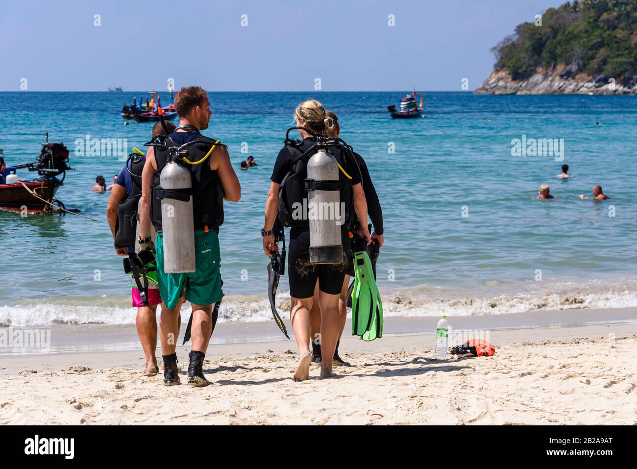 Vier Taucher mit Sporttanks gehen in Richtung Meer am Kata Beach, Phuket, Thailand Stockfoto