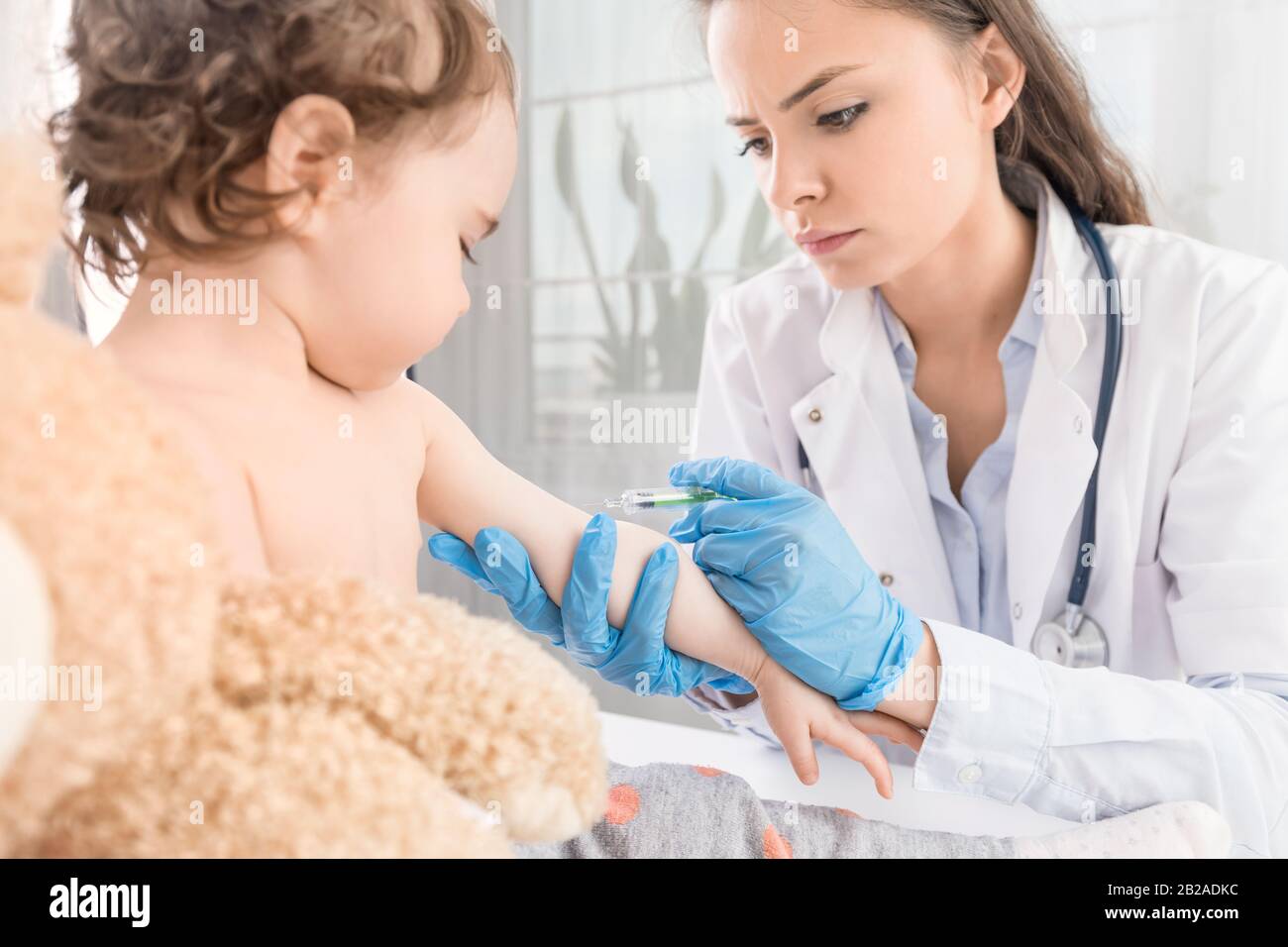 Junge Frau Kinderärztin führt eine Impfung eines kleinen Mädchens durch. Das Mädchen hält ein Maskottchen. Stockfoto