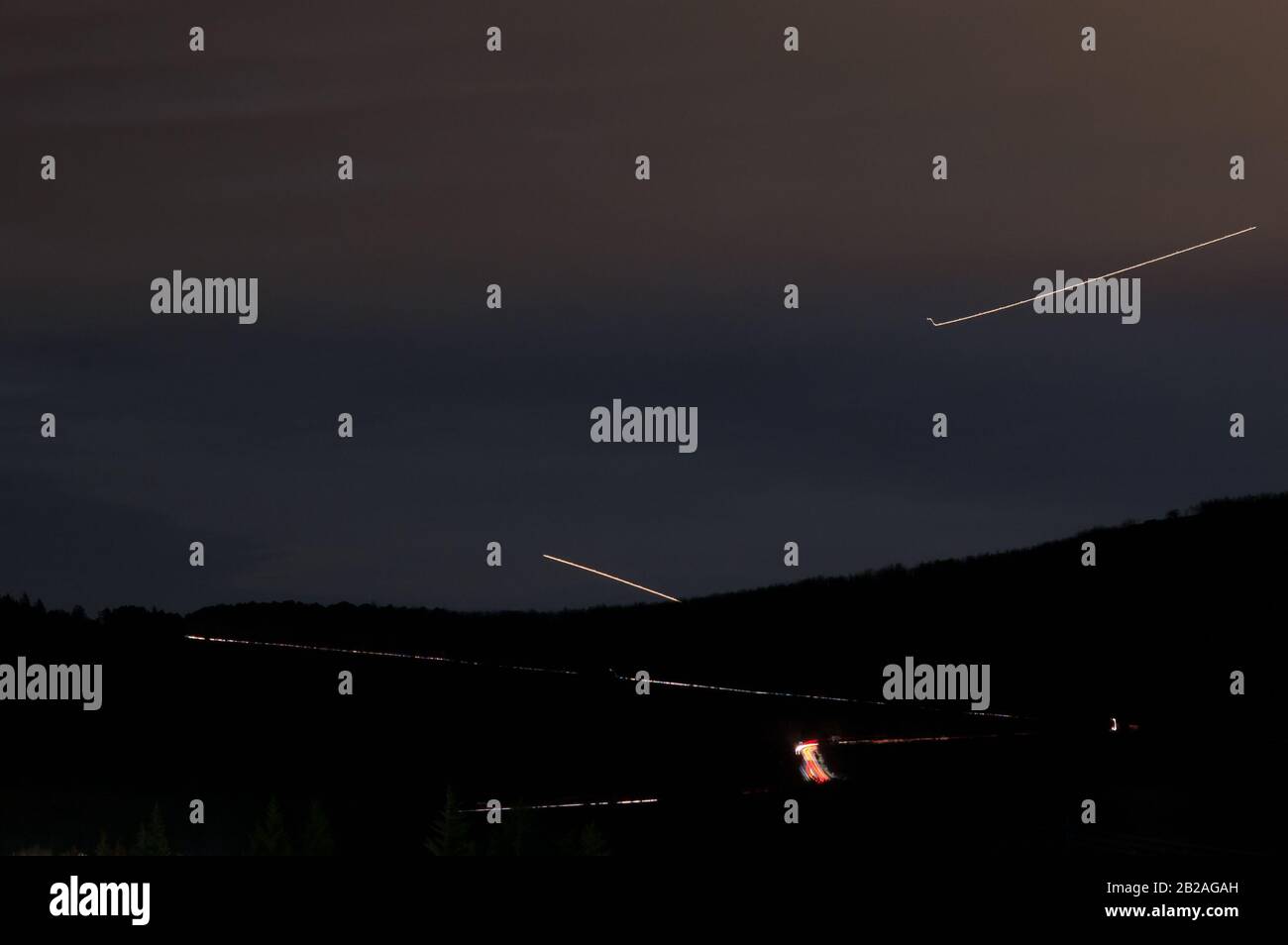 Lange Belichtungszeit mit Lichtwegen einer Straße und Lichtwegen von zwei Flugzeugen mit dem Berg in Silhouette Stockfoto