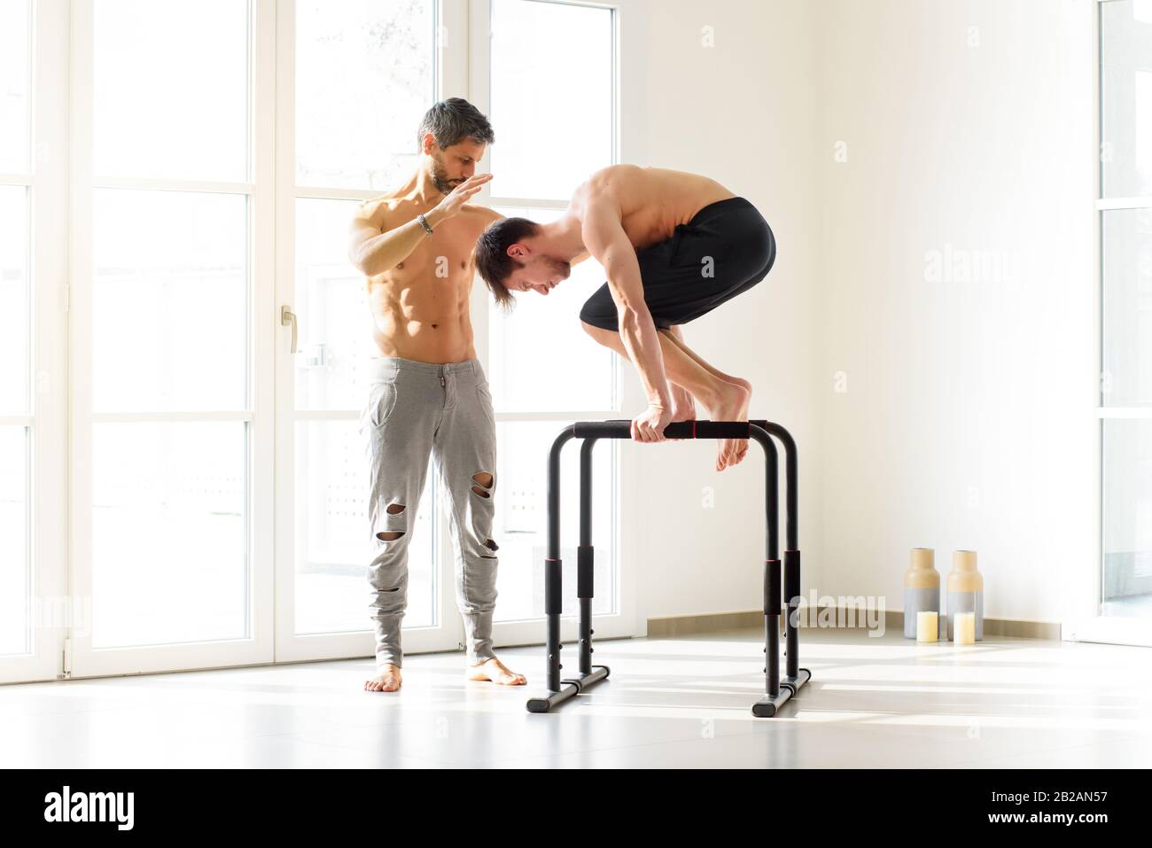 Junger Mann, der eine planche Kalisthenik-Übung auf parallelen Stangen macht, unterstützt von einem fit muskulösen Personal Trainer in einer hellen High-Key-Turnhalle mit Kopierraum Stockfoto