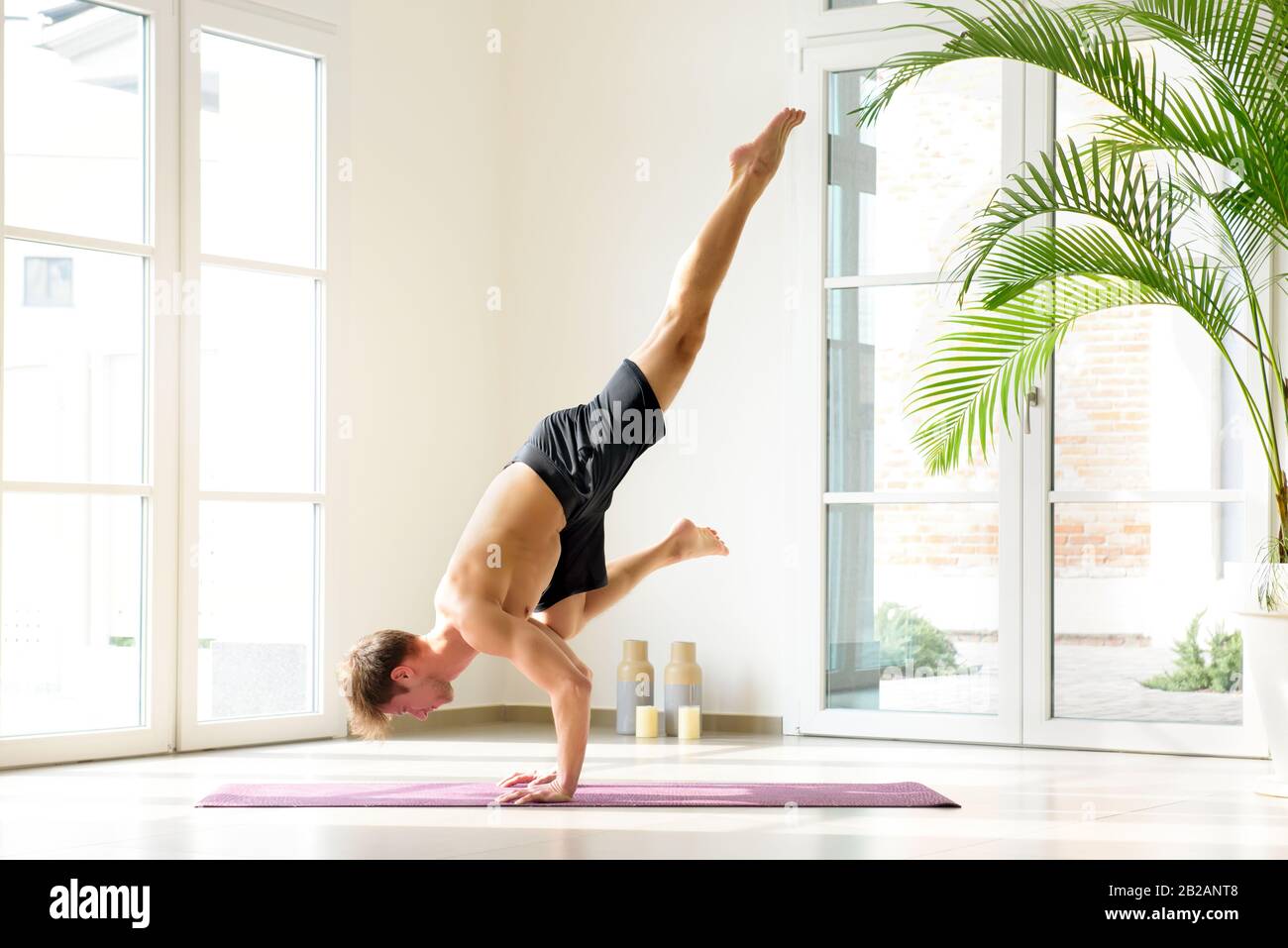 Muskulöser junger Mann, der Yoga in Flying Crow macht und drinnen gegen helle Fenster und weiße Wände arbeitet Stockfoto