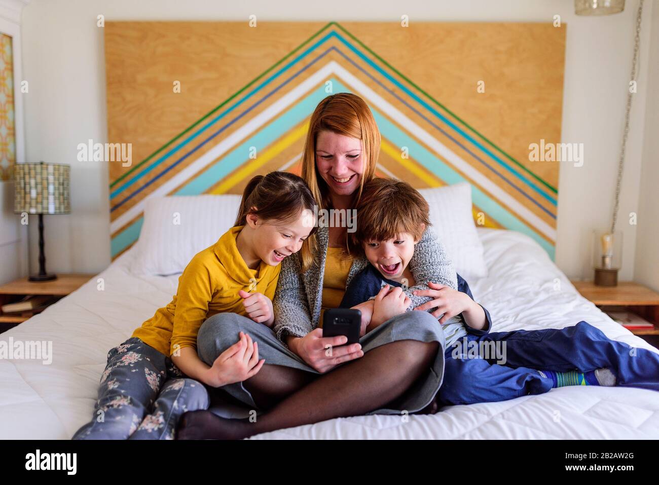 Fröhliche Mutter sitzt mit zwei Kindern auf dem Bett und schaut auf ein Handy Stockfoto
