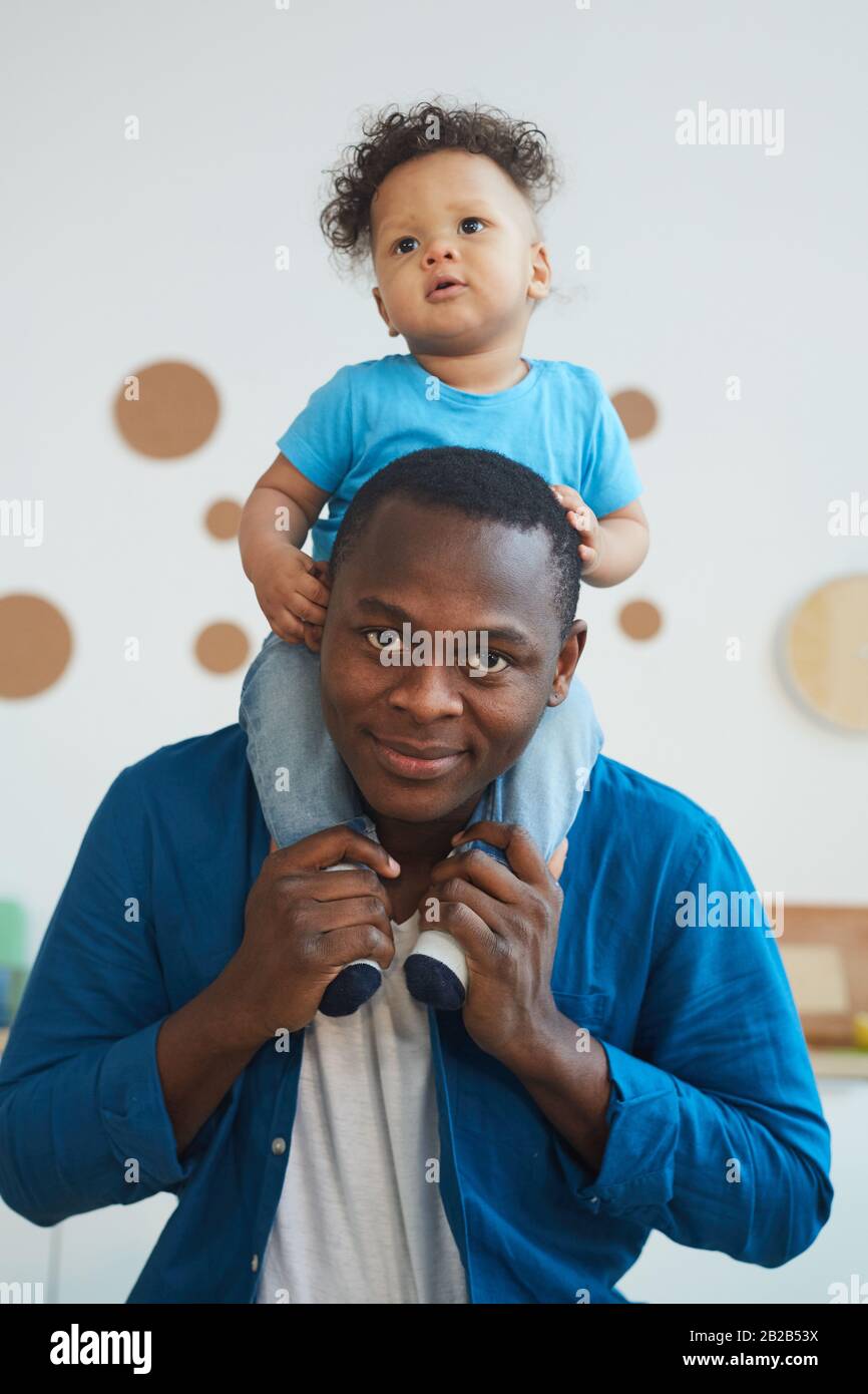 Vertikales Porträt eines glücklichen afroamerikanischen Mannes, der Sohn auf den Schultern trägt und lächelnd mit der Kamera blickt Stockfoto