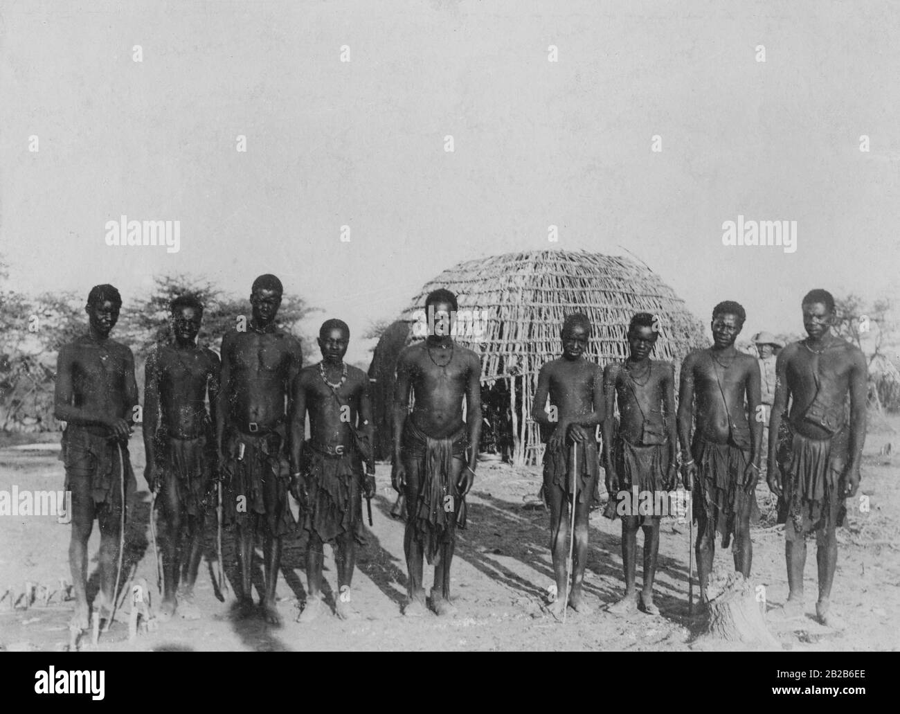 Eine Gruppe von Männern aus Herereo in einem Dorf. Im Hintergrund ist Mitglied der Deutschen Schutztruppe. Undatiertes Foto. Stockfoto