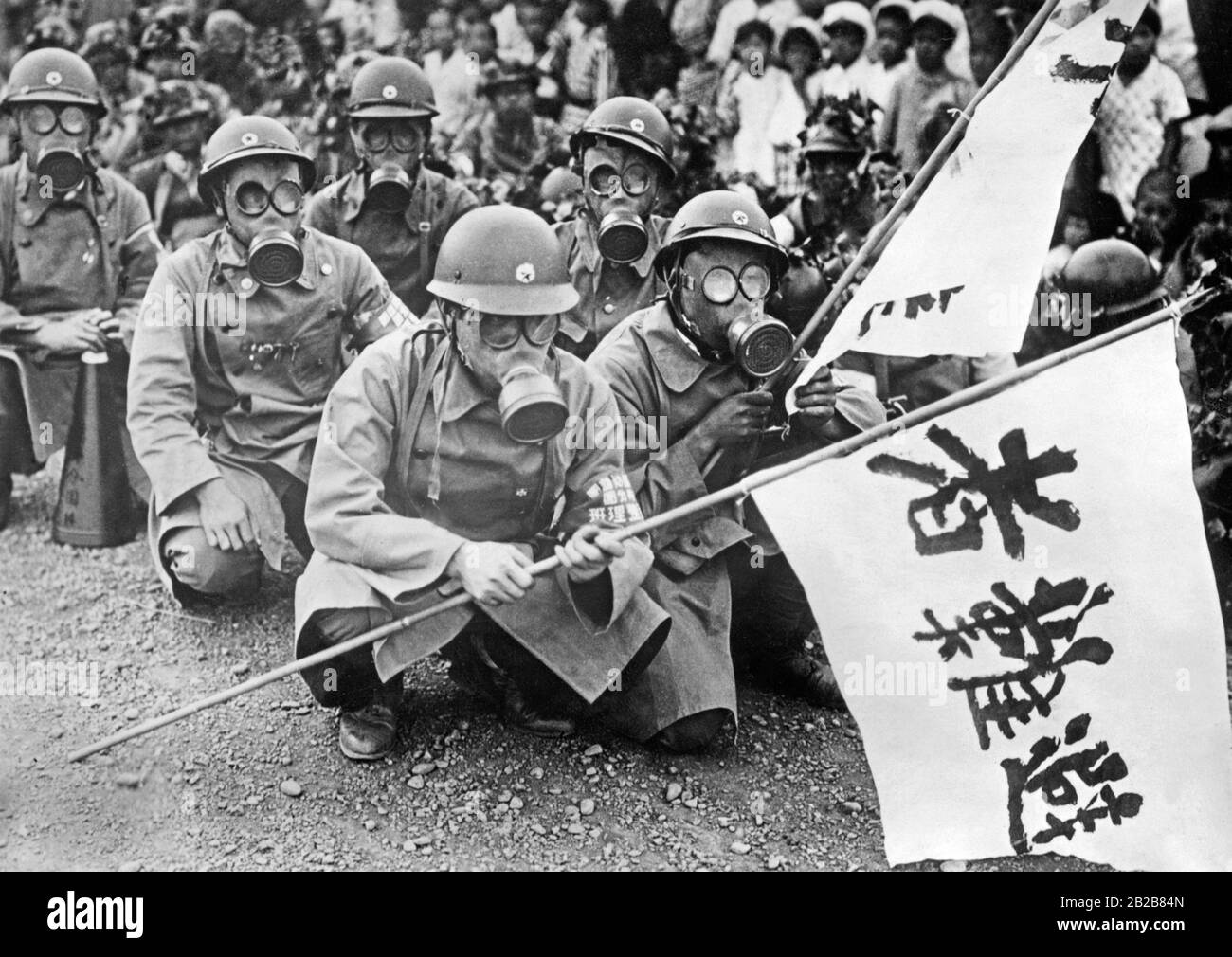 Eine Gruppe japanischer Soldaten quietschte während einer luftangriffübung auf einer Straße in einer japanischen Stadt. Sie tragen Gasmasken und Fahnen. Im Hintergrund Zuschauer aus der Stadt. Stockfoto