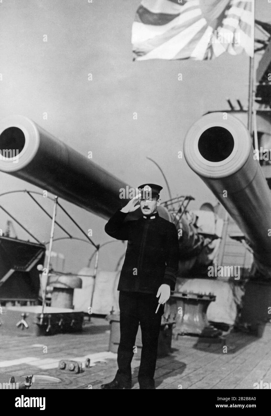 VizeAdmiral der Kaiserlich japanischen Marine an Deck eines japanischen Kriegsschiffs. Er salutiert vor einer großen Kanone. Im Hintergrund die Flagge der japanischen Marine. Stockfoto
