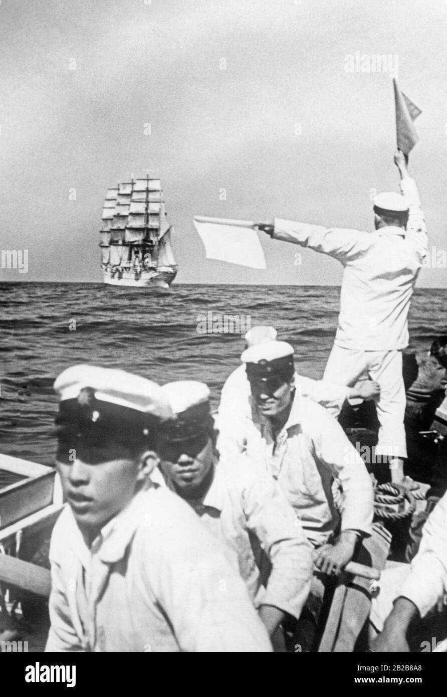 Blick auf das Meer vor der japanischen Küste. Im Vordergrund stehen einige Kadetten der Marineschule Tokio der Kaiserlich japanischen Marine in einem Ruderboot. Einer von ihnen hält mittels Signalfahnen Kontakt zu ihrem Ausbildungsschiff, der Taisei Maru. Stockfoto