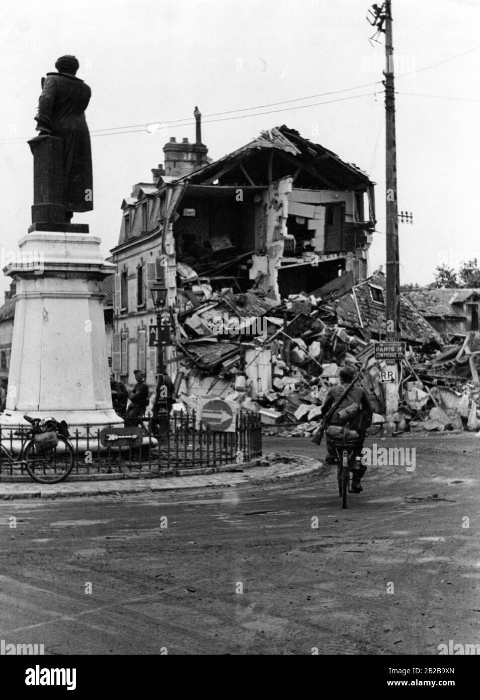 Ein deutsches Rad-Bataillon, das auf dem Weg nach Paris eine französische Stadt durchquert. Im Hintergrund steht ein im Krieg zerstörtes Haus. Auf der linken Seite befindet sich ein unversehrtes Denkmal. Stockfoto