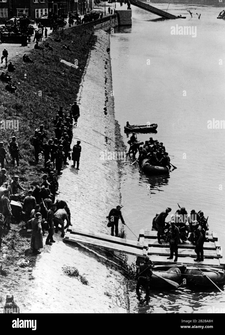 Einmarsch in Holland 1940: Das Bild zeigt Fahrzeuge und Soldaten, die mit Booten die Maas überqueren und im Rücken die zerstörte Wilhelminen-Brücke. Stockfoto
