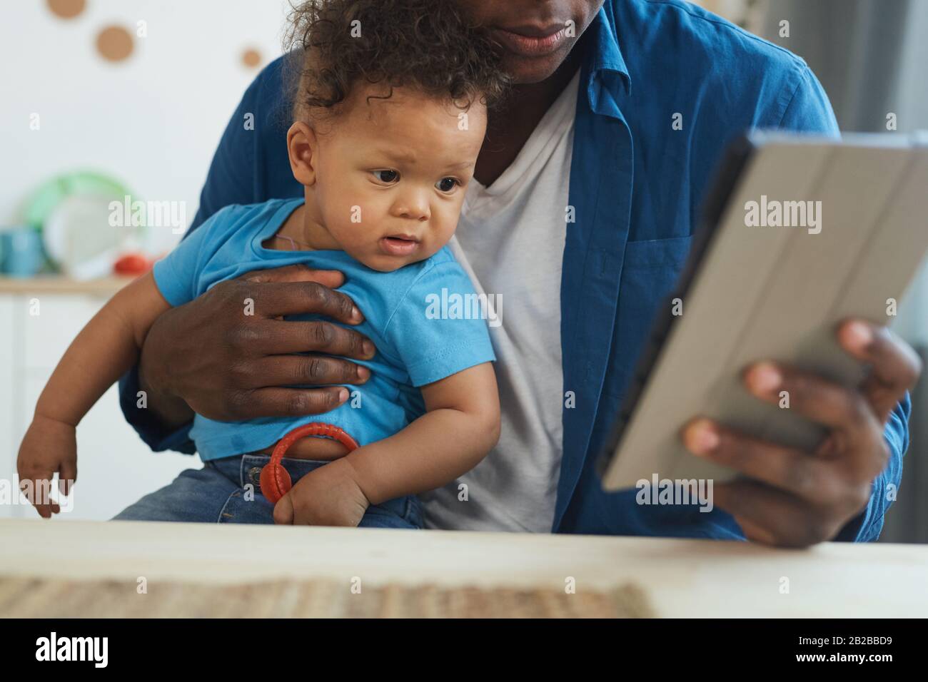 Zugeschnittenes Porträt eines afroamerikanischen Mannes, der Baby hält, während er ein digitales Tablet im Innenbereich verwendet, Platz für Kopien Stockfoto
