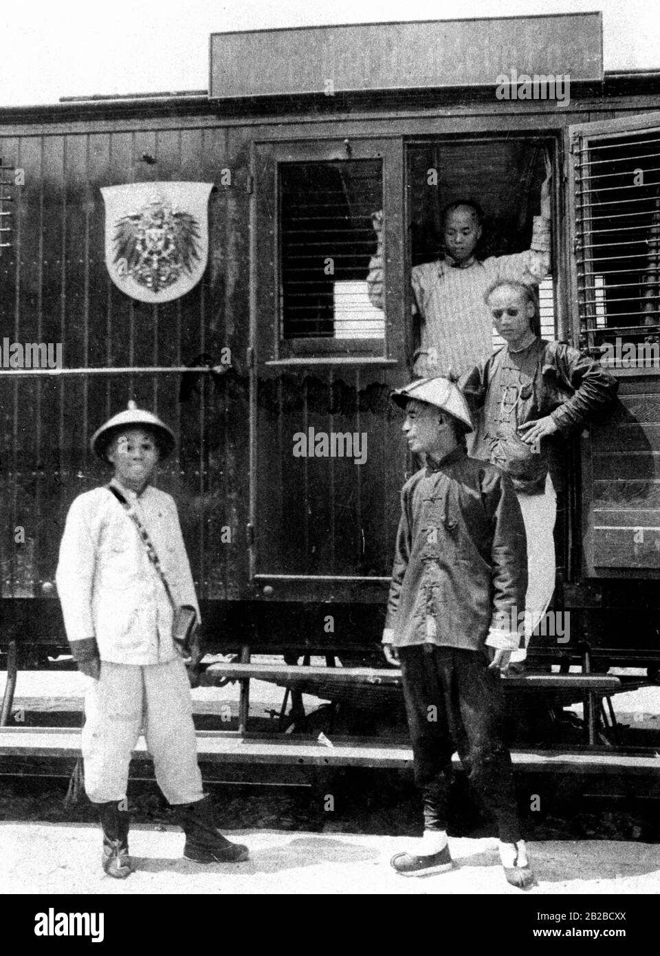 Chinesische Beamte der kaiserlichen, deutschen Reichspost auf der Shantung-Eisenbahnlinie in Tsingtau vor ihrem Postwagen, der den schwarzen "Hoheitsadler" (Nationaladler) des Deutschen Kaiserreichs trägt. Stockfoto