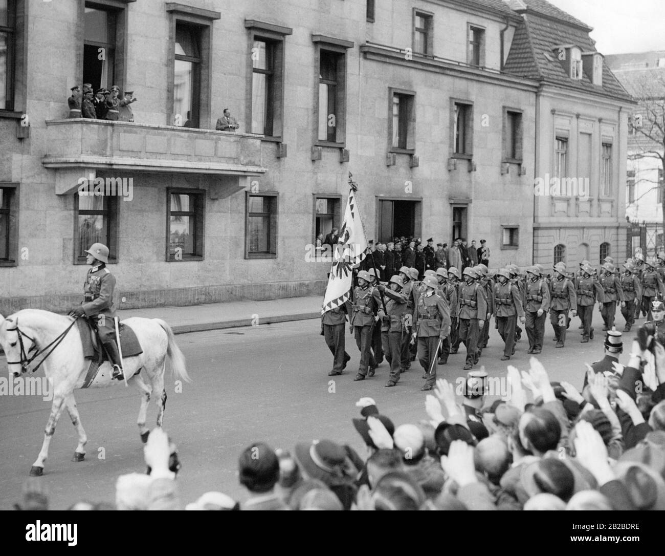 Das österreichische Bataillon marschiert an der neuen Staatskanzlei vorbei. Adolf Hitler steht auf dem Balkon und macht den Nazi-Salut. Der führende Soldat trägt die Flagge der Kaiserlichen Kaiserlichen Kaiserlichen Trabantgarden. Sie werden von Passanten am Straßenrand mit dem Nazi-Salut begrüßt. SS-Männer stehen an einem Seiteneingang der Staatskanzlei. Stockfoto