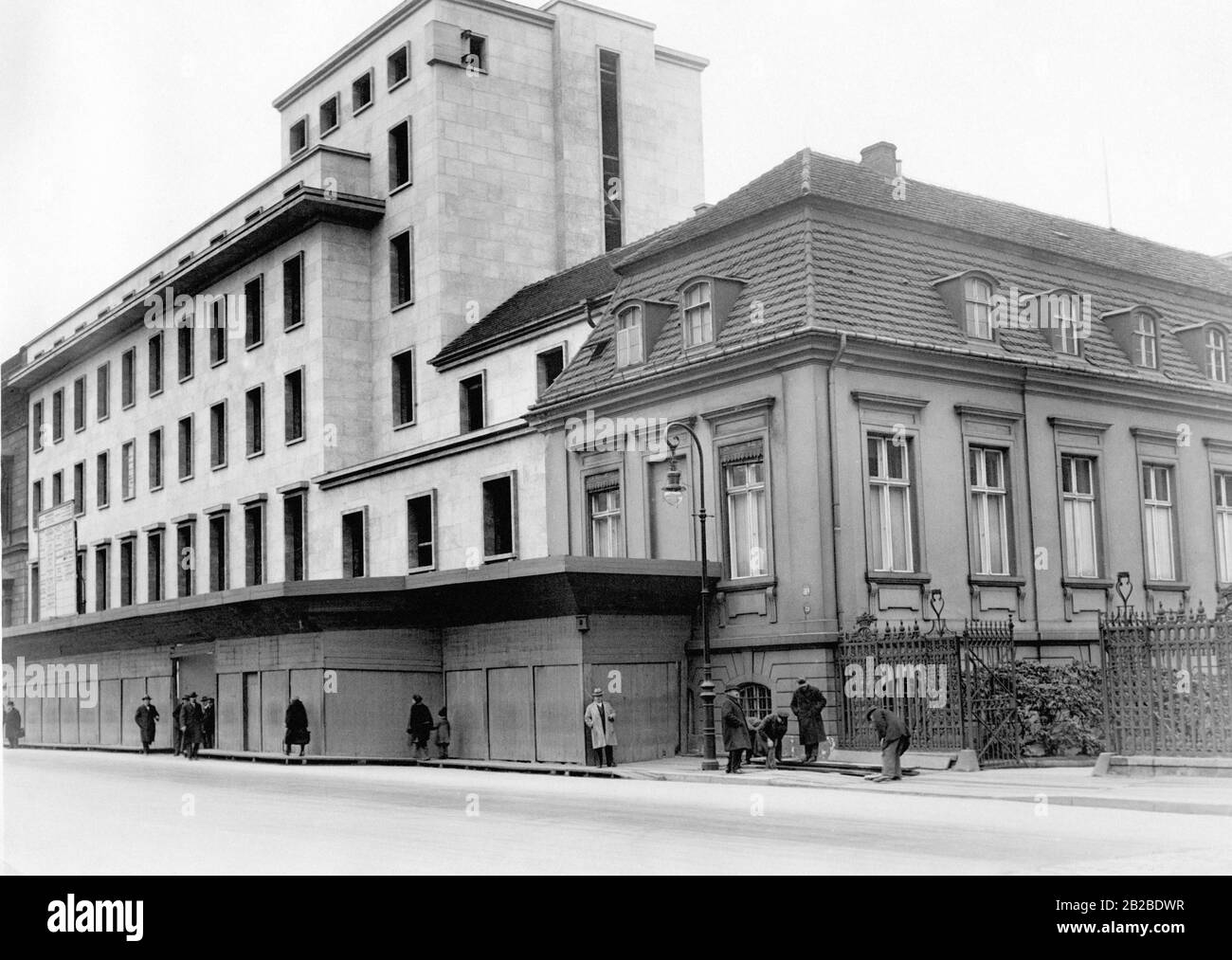 Das Bild zeigt die neuen und Teile der alten Reichskanzlei von vorne. Die Menschen verlassen das Gebäude, Handwerker bauen rechts auf dem Bürgersteig eine Rampe. Stockfoto