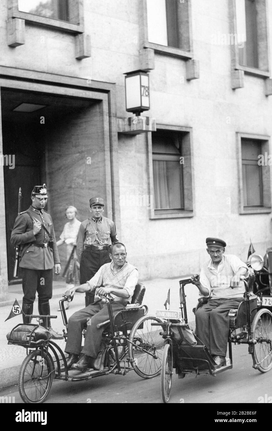 Zwei Kriegsinvaliden mit ihrem Dreirad vor der Staatskanzlei. Sie fuhren von Hindenburgs in Obermösien nach Berlin, um von Bundeskanzler Adolf Hitler empfangen zu werden. Ein Polizist und ein SS-Mann bewachen den Eingang. Stockfoto