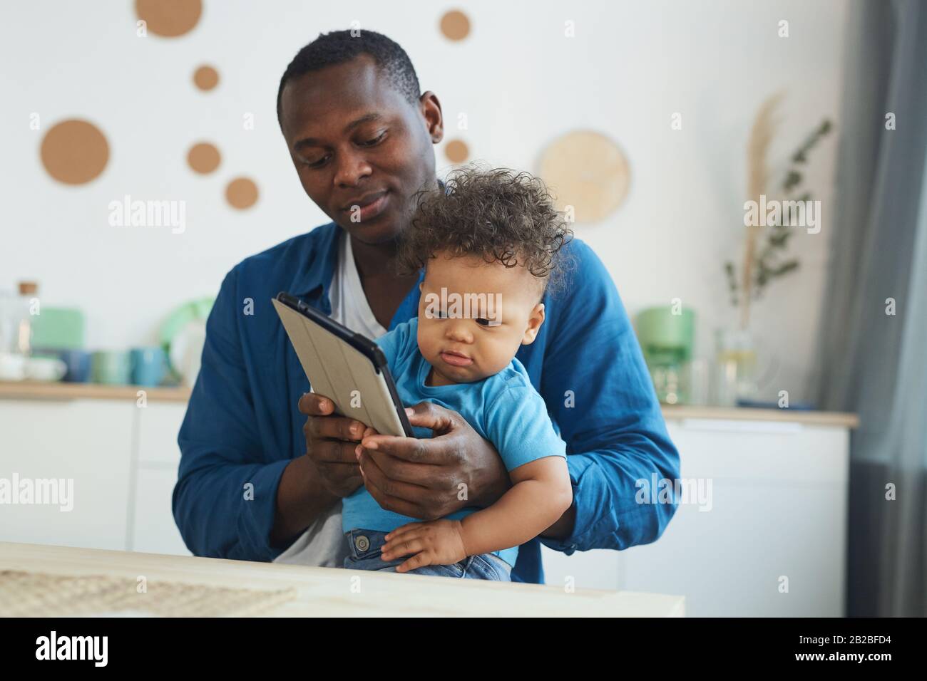 Porträt eines afroamerikanischen Mannes, der weinendes Baby hält, während er digitales Tablet im Innenbereich verwendet, Platz für Kopien Stockfoto