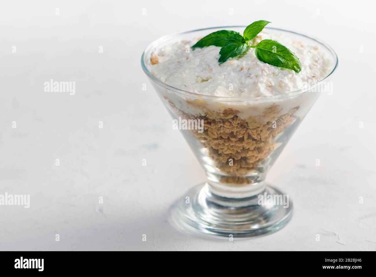 Granola in einer Schüssel mit Joghurt und Basilikumblättern. Granatschüssel auf strukturiertem weißem Tisch. Stockfoto