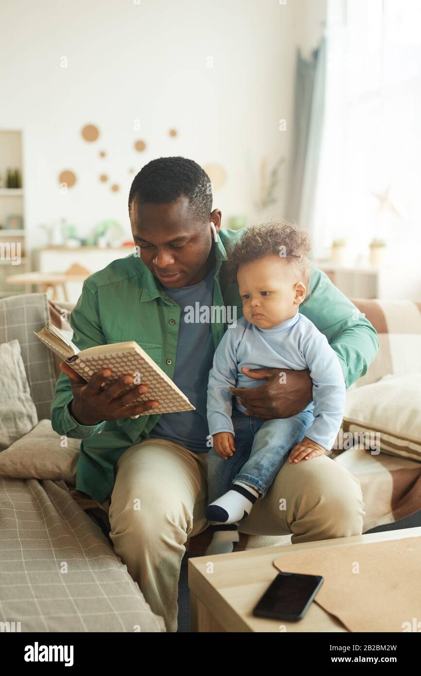 Vertikales Porträt eines reifen afroamerikanischen Mannes, der Buch zu Kind liest, das auf dem Schoß der Väter sitzt Stockfoto